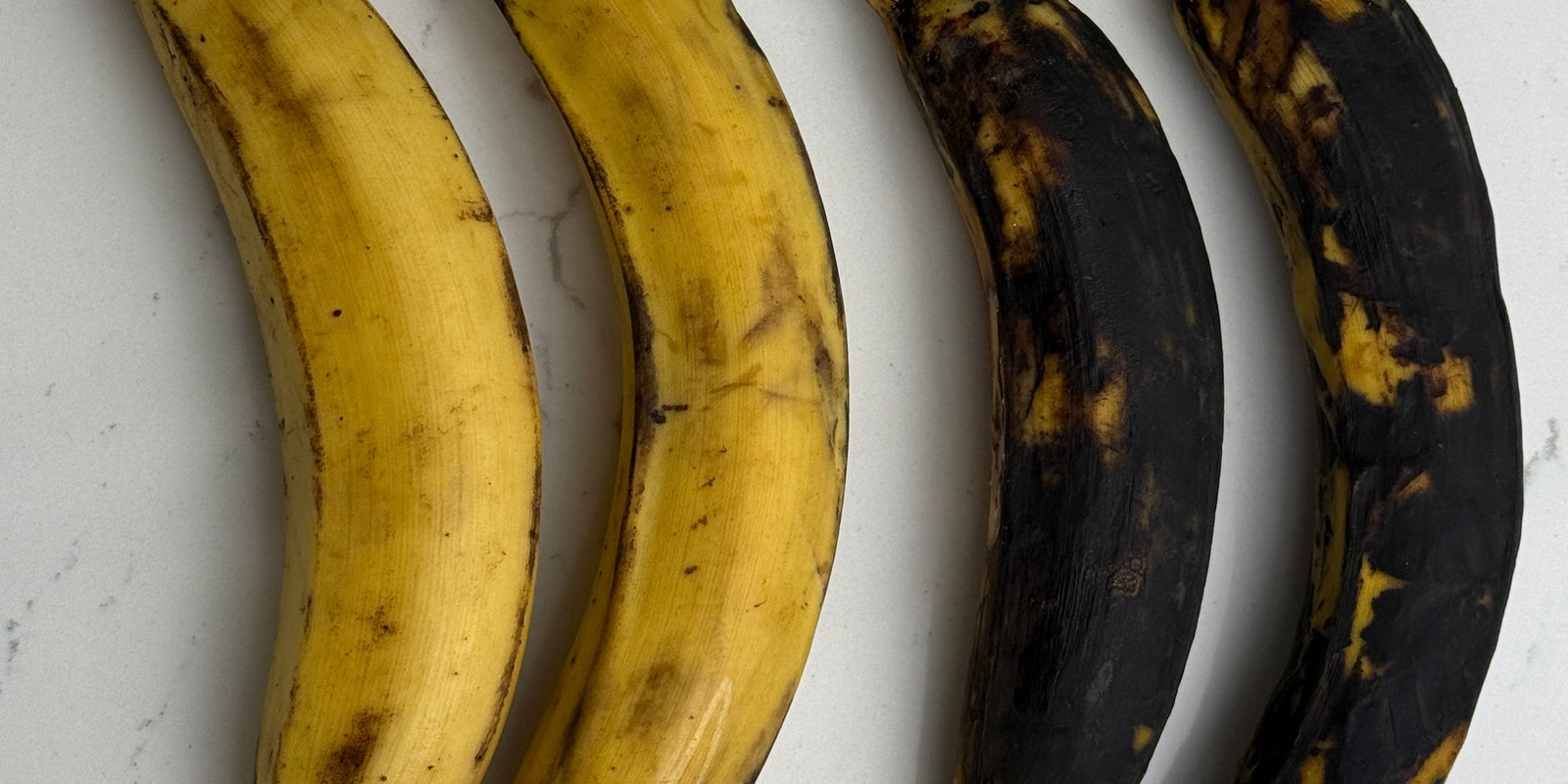  Four plantains displayed on a white countertop showing different ripeness stages — two very ripe with black skins on the left and two yellow, less ripe plantains on the right.